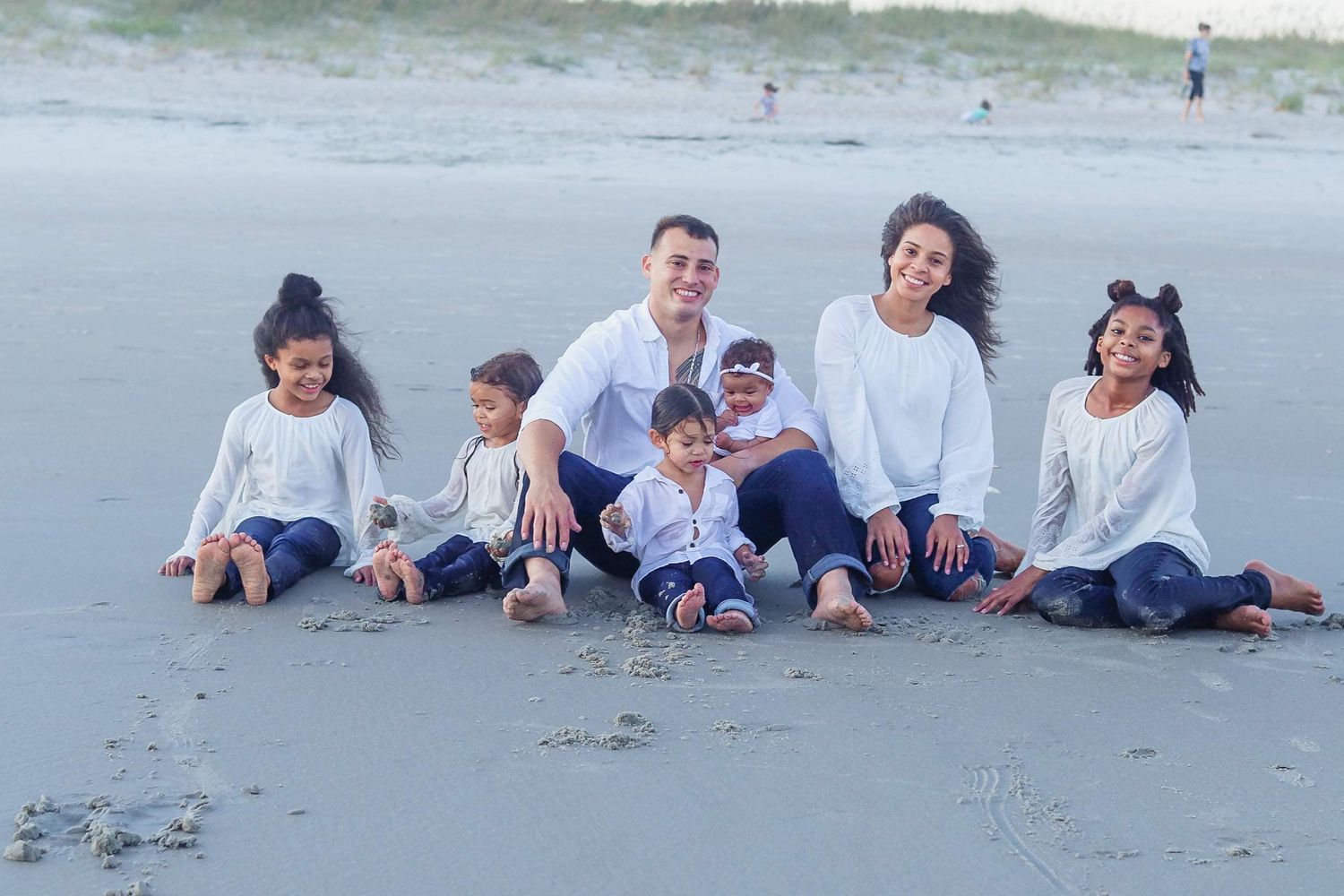Happy family of seven poses together on a sandy beach in matching white outfits.