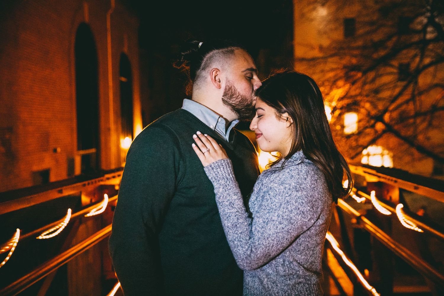Romantic couple shares a tender moment together on a bridge lit by warm golden lights at night.