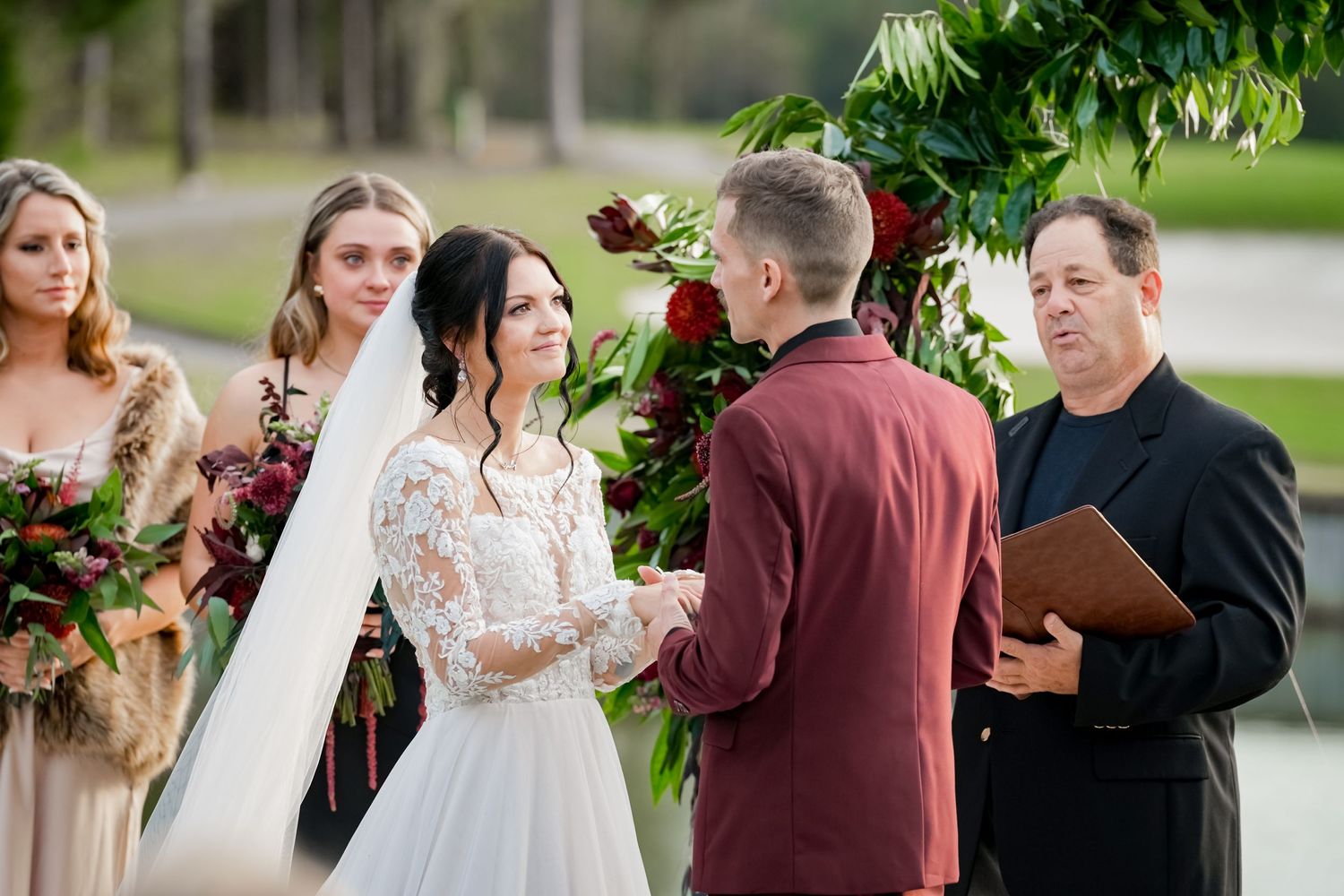 Outdoor wedding ceremony under greenery arch with wedding party and officiant.