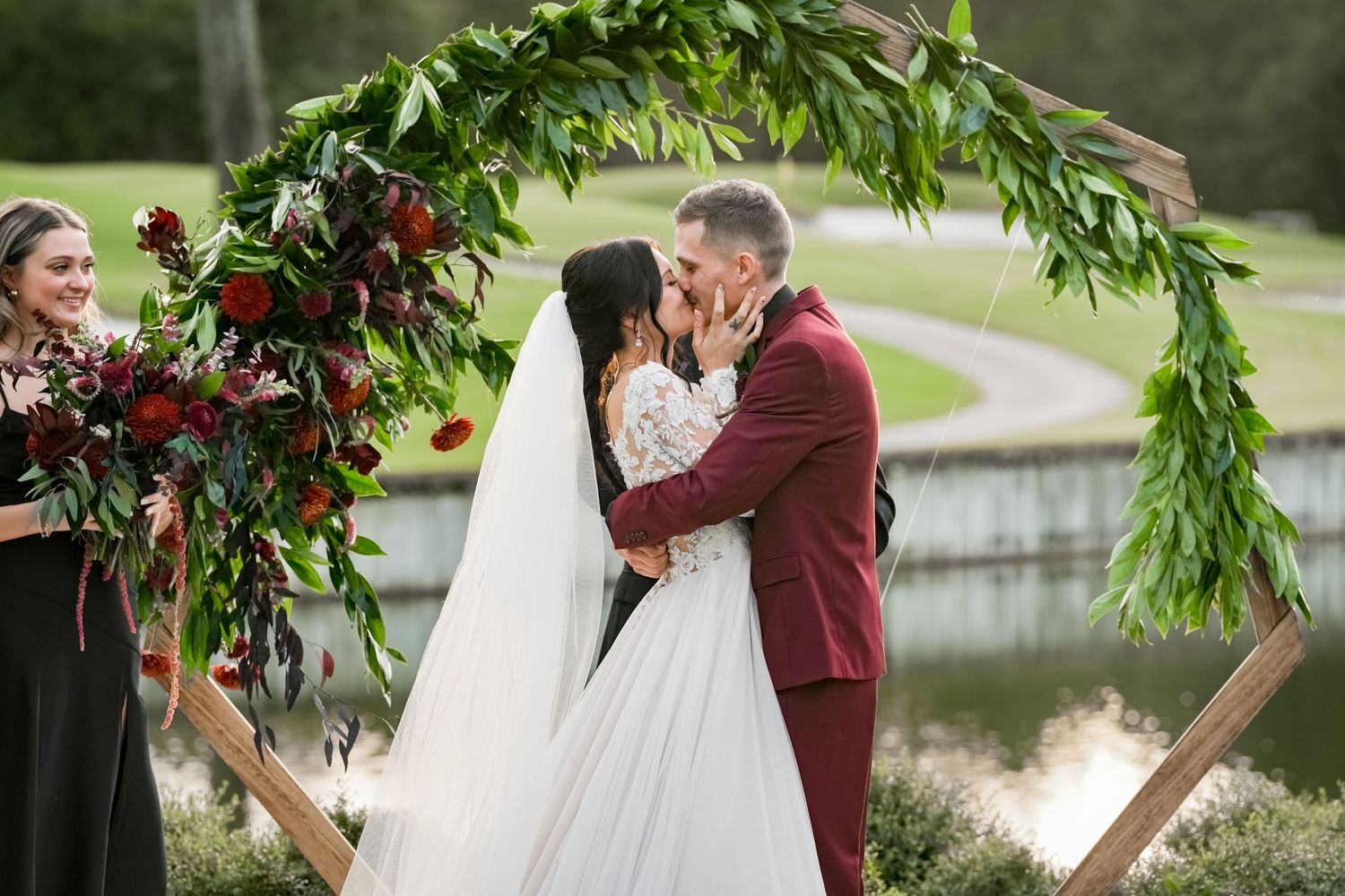 First kiss as newlyweds under floral arch with burgundy flowers and greenery.