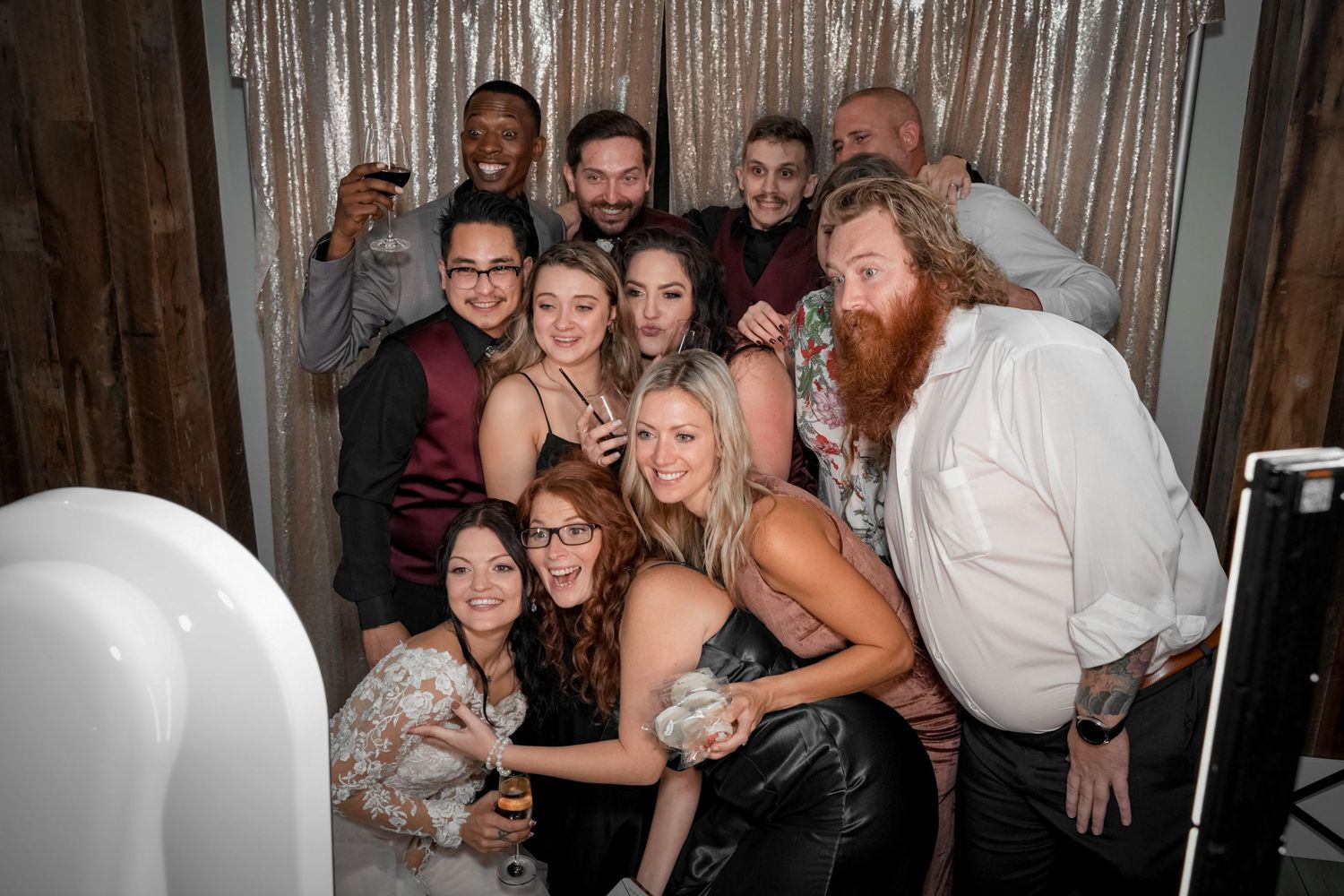 Large group celebrating at wedding reception with photo booth and sparkly backdrop.