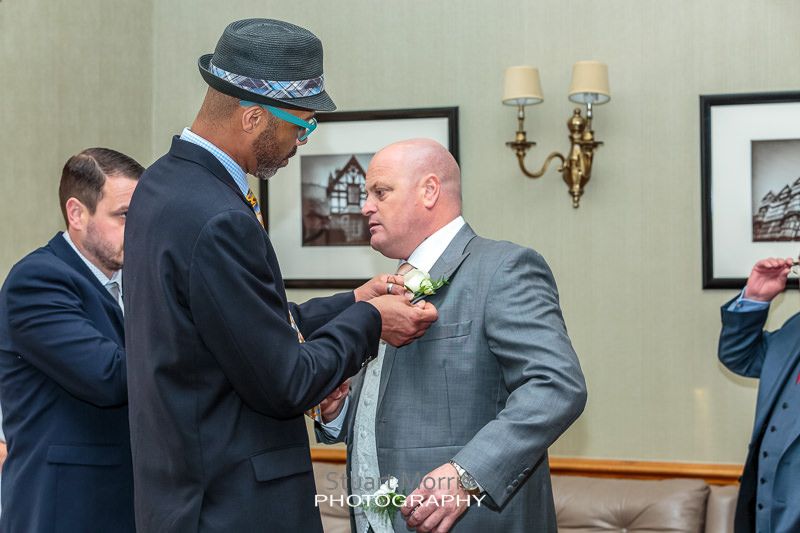 wedding guest wearing a trilby helps the groom attach his button hole to his lapel
