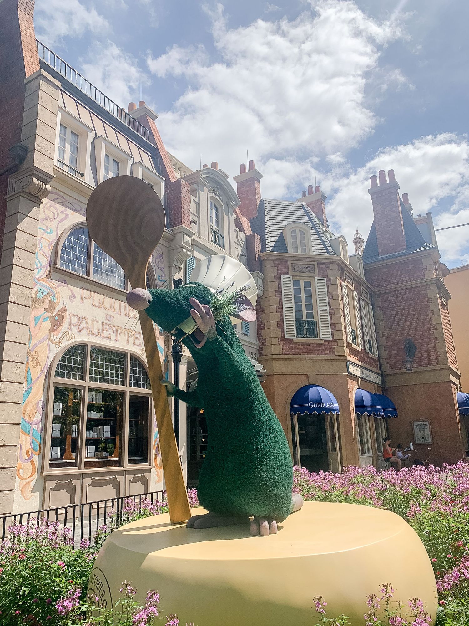 Green topiary sculptures displayed along a garden path with European-style buildings in background.