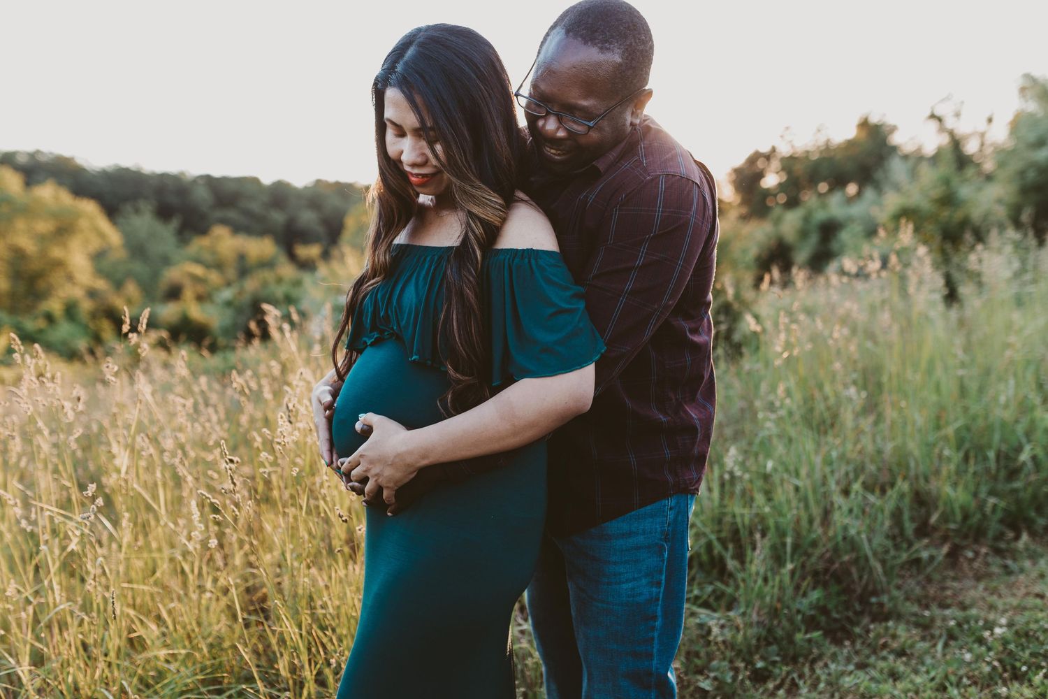Woman wearing maternity gown smiles as her husband hugs her