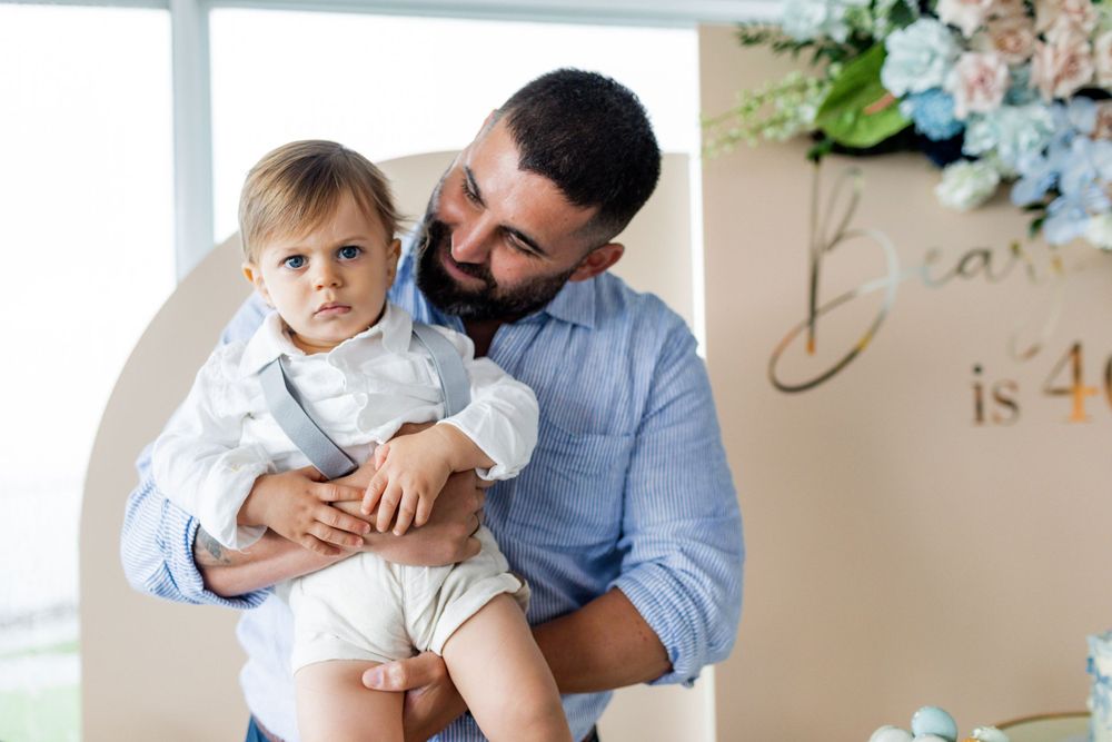 Baby dressed in white sits on parent's lap by window during first birthday celebration.