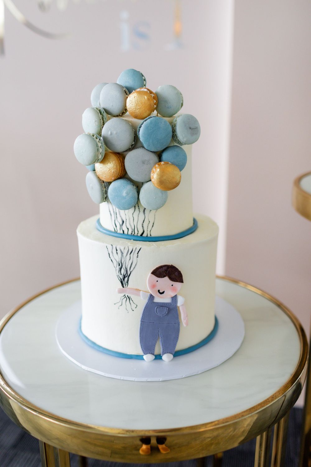 White birthday cake decorated with blue and gold macarons arranged like balloons with a cute illustrated figure.