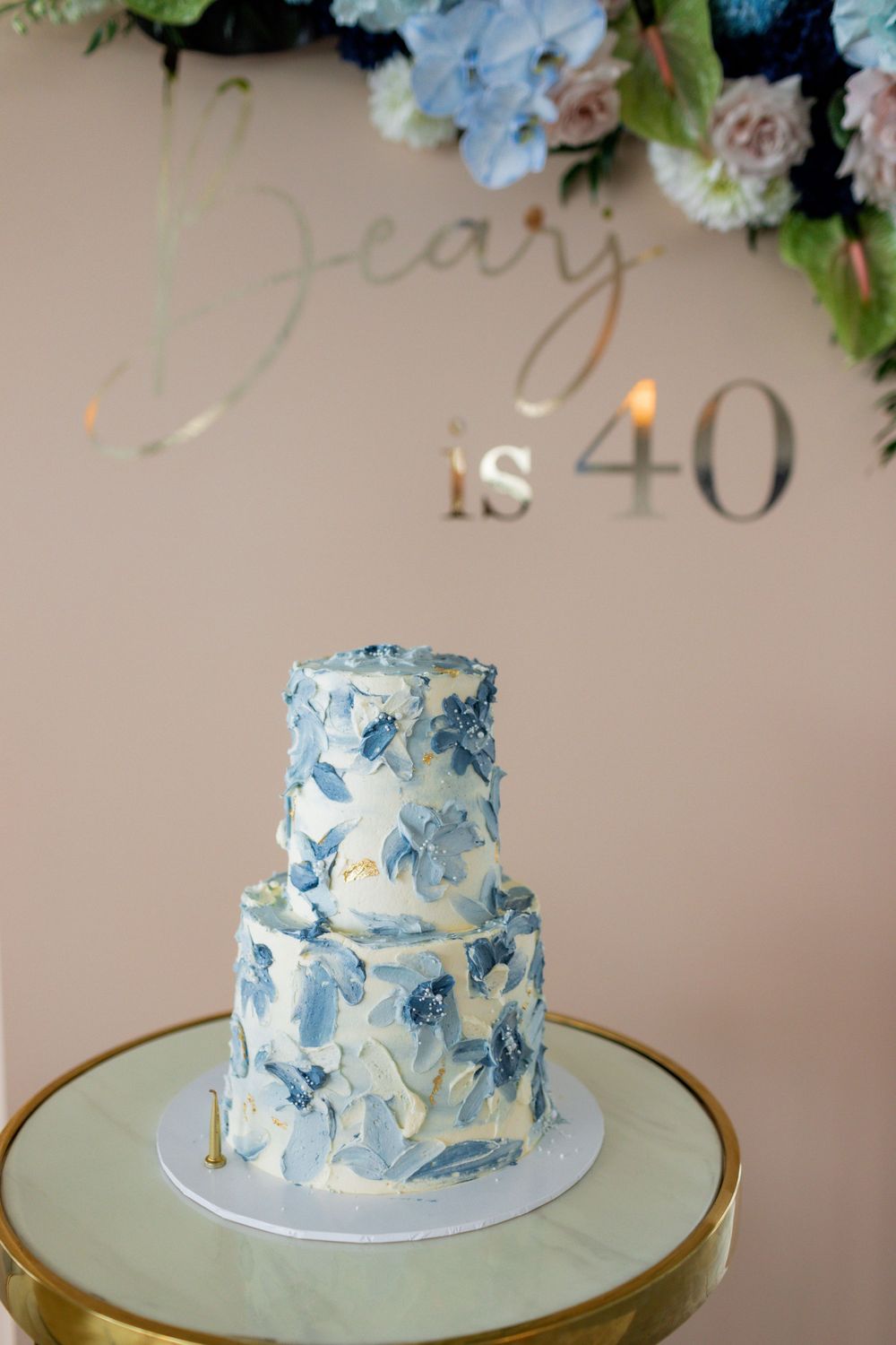 Two-tiered white cake with delicate blue floral designs displayed on a gold pedestal.