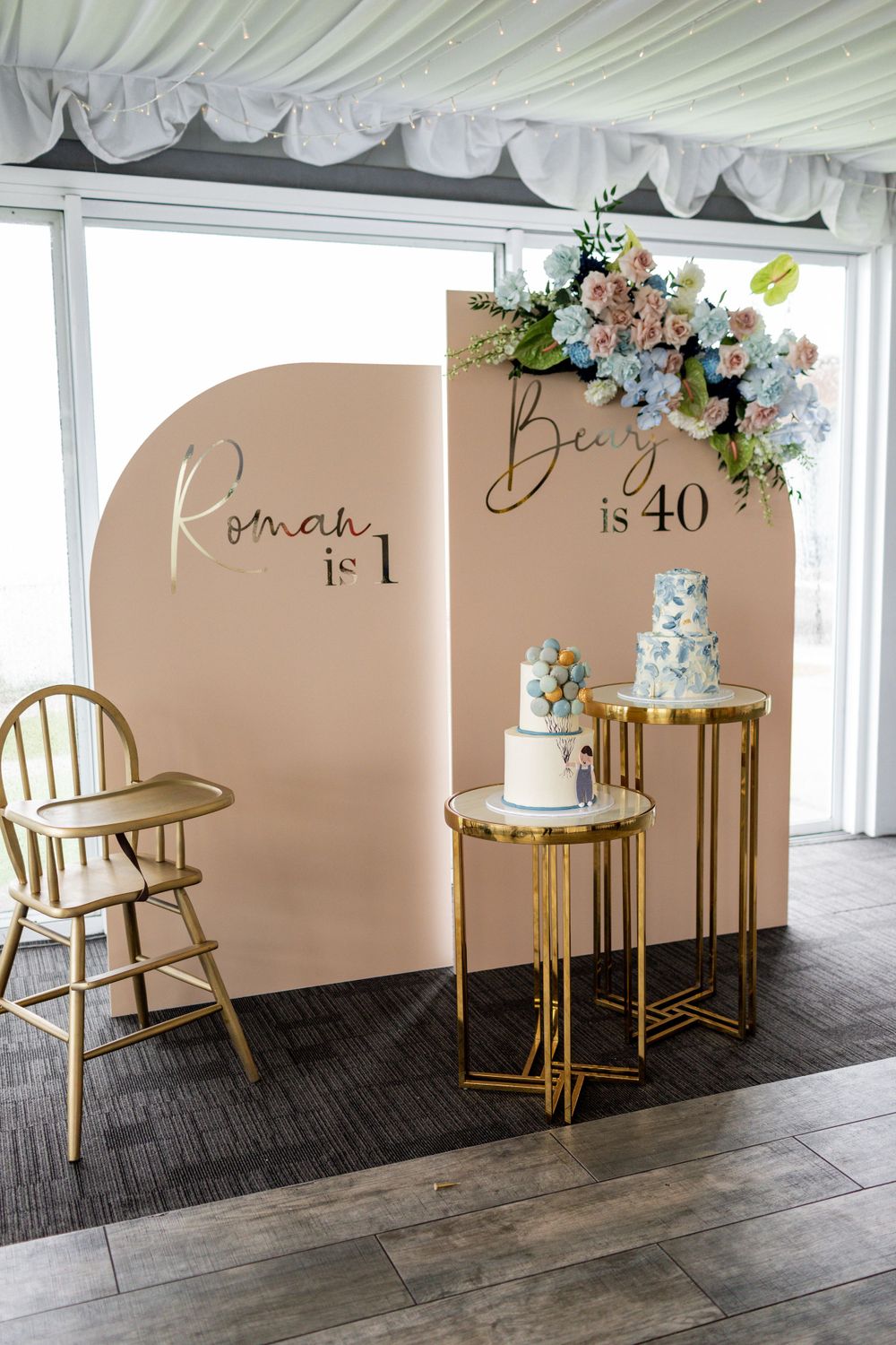 Modern party display featuring tiered cakes on gold stands against a blush backdrop with floral accents.