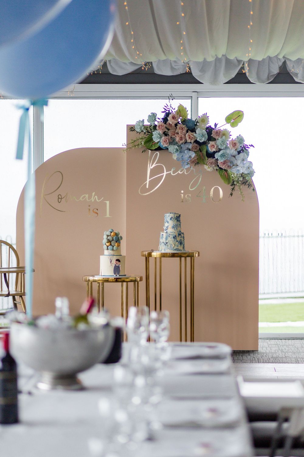 Party setup with tiered cakes on gold pedestals against a beige backdrop with floral decorations.