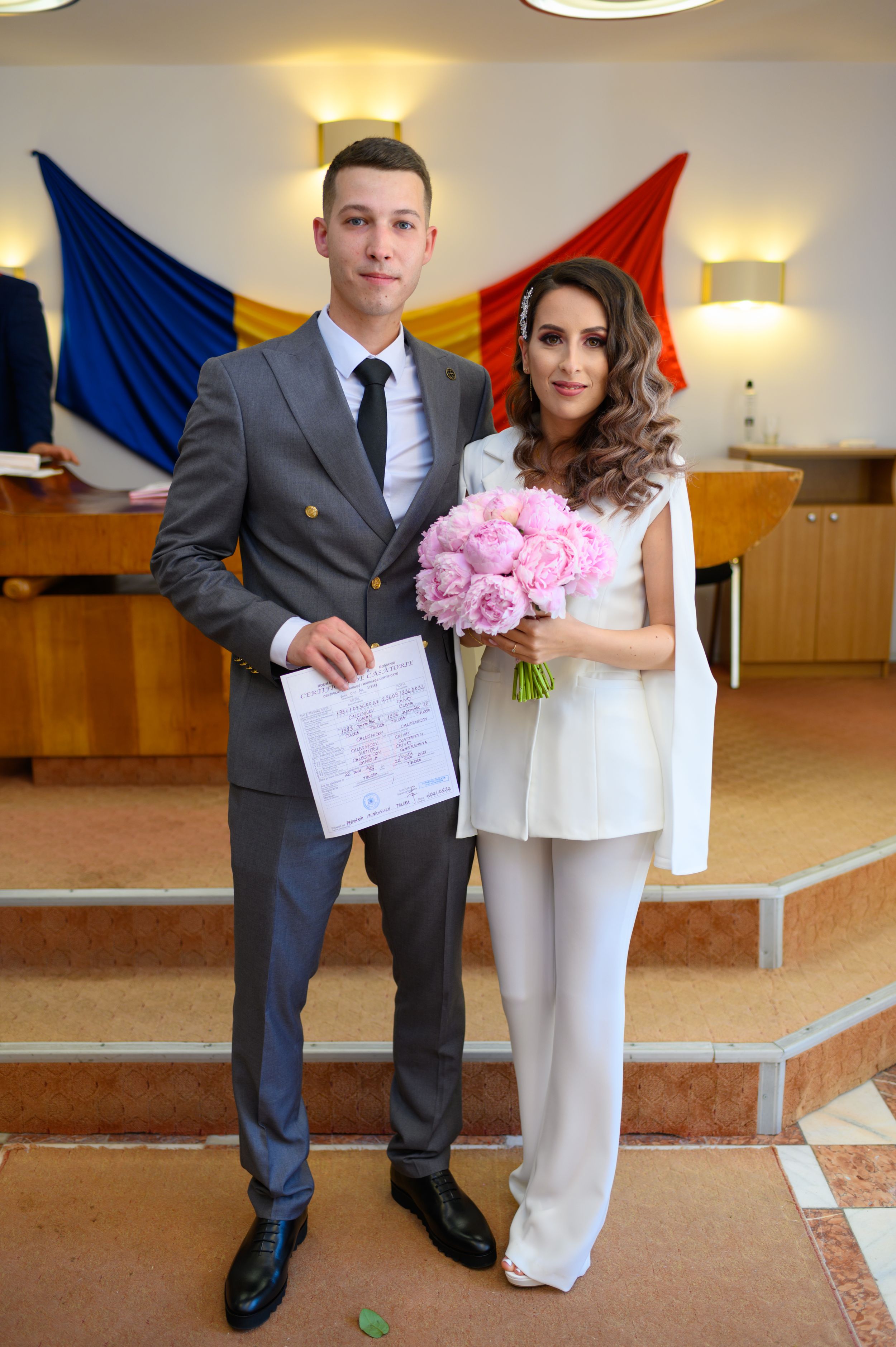 A couple in formal attire posing with a pink bouquet in front of a tricolor flag.
