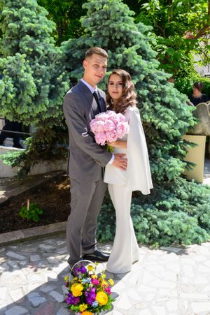 A couple in formal attire posing with pink flowers against evergreen trees.