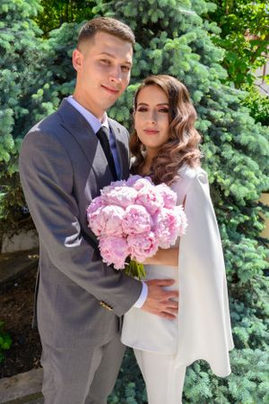 A couple in grey suit and white outfit holding pink flowers in a garden setting.