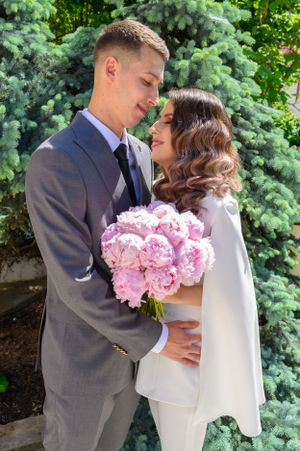An intimate moment between newlyweds with pink peonies against evergreen trees.