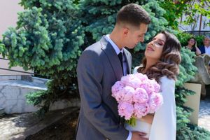 Elegant couple embracing outdoors with pink floral bouquet and garden backdrop.