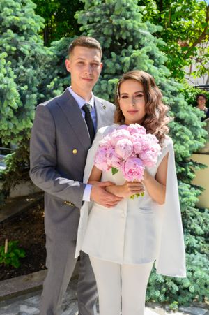 Wedding couple in grey suit and white dress hold pink peony bouquet in garden setting.