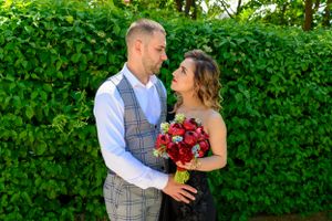 Couple in formal wear share intimate moment with red roses against green foliage.