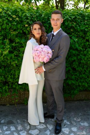 Couple in grey suit and white outfit pose with pink flowers against garden wall.
