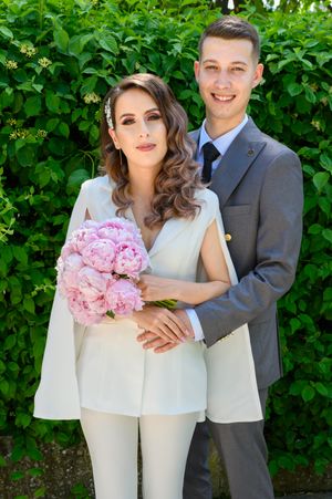 Bride in white pantsuit holds pink peonies while posing with groom by green hedge.