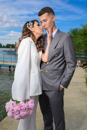 A romantic wedding couple embraces by a lake, with the bride holding pink peonies and wearing a white cape dress.
