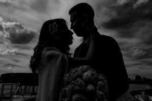 A silhouetted couple shares an intimate moment against a dramatic cloudy sky by the water.