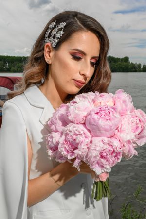 A bride in white holds a lush bouquet of pink peonies by a lake, wearing an elegant hair accessory.