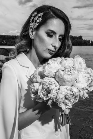 A black and white portrait of a bride holding a peony bouquet near a lake.