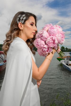 A bride in a flowing white cape dress admires her pink peony bouquet by the waterfront.