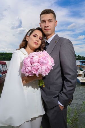 A wedding couple poses by the water, with the bride in white holding pink peonies.