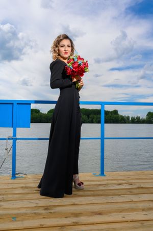 A woman in a black formal dress holds red roses while posing on a dock by the water.