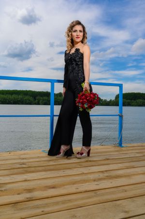 A woman in an elegant black jumpsuit poses with red roses on a lakeside dock.