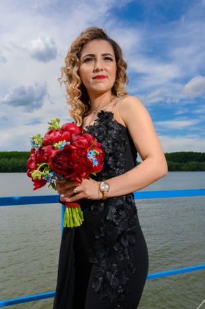 A woman in a black lace dress holds a vibrant red rose bouquet on a waterfront deck.
