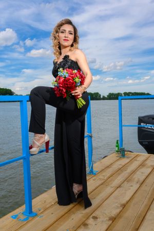 A woman in a black formal outfit poses with red roses on a wooden dock by the lake.