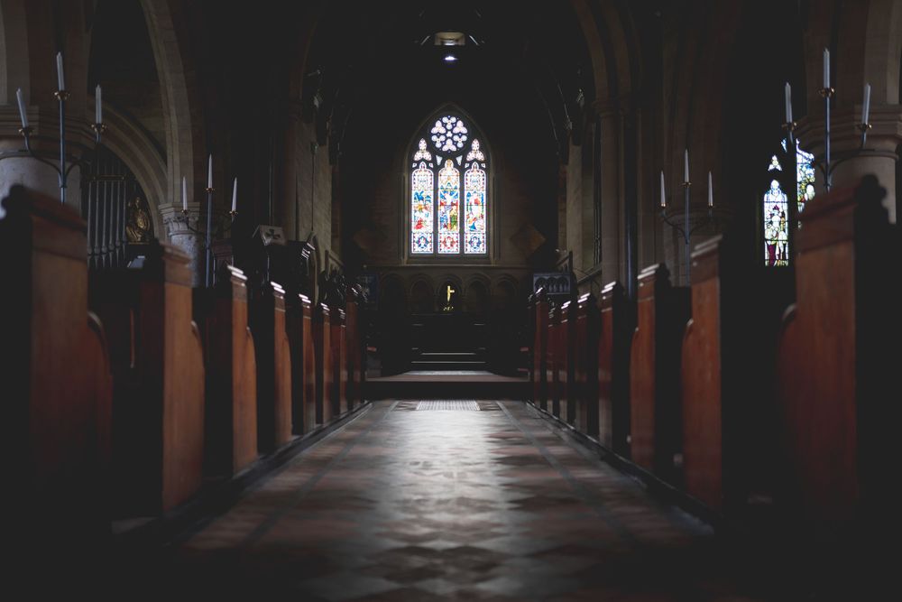 Dark church interior with stained glass windows and wooden pews creating dramatic lighting.