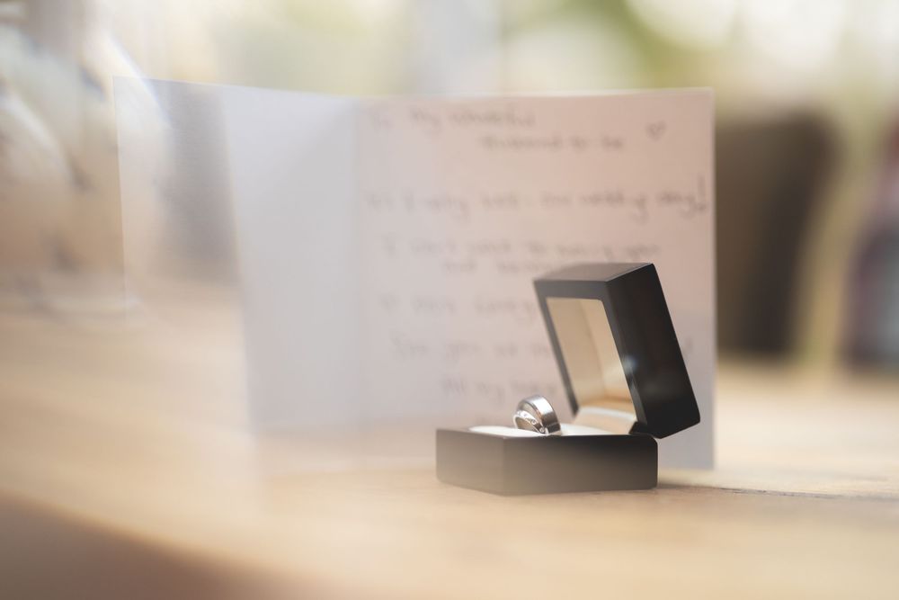 An engagement ring box sits on a blurred surface with a handwritten note in the background.