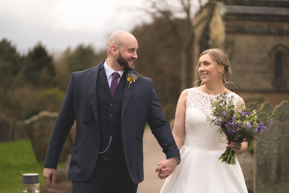 A newlywed couple walks together outdoors holding hands with a church visible in the background.