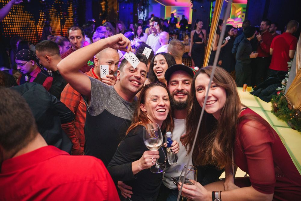 Party scene with people taking selfies and raising drinks in celebration at a nightclub.