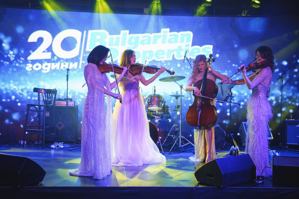 Musical performers in white dresses playing string instruments on stage with blue backlighting.