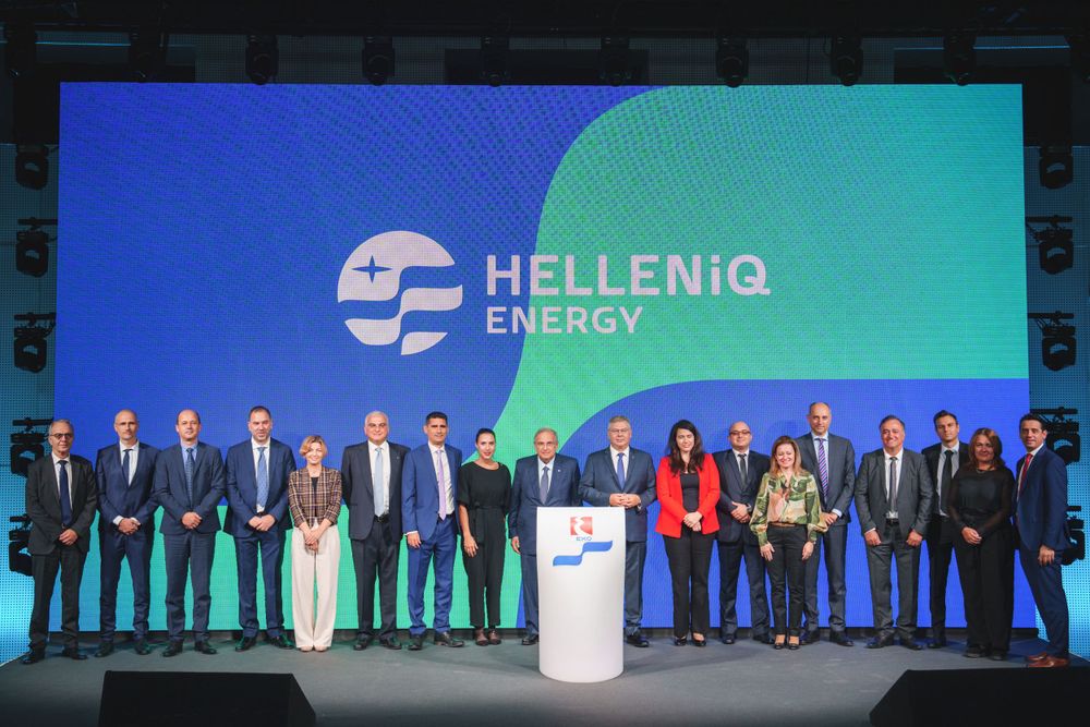 Business professionals stand on stage at Helleniq Energy corporate event with branded backdrop.