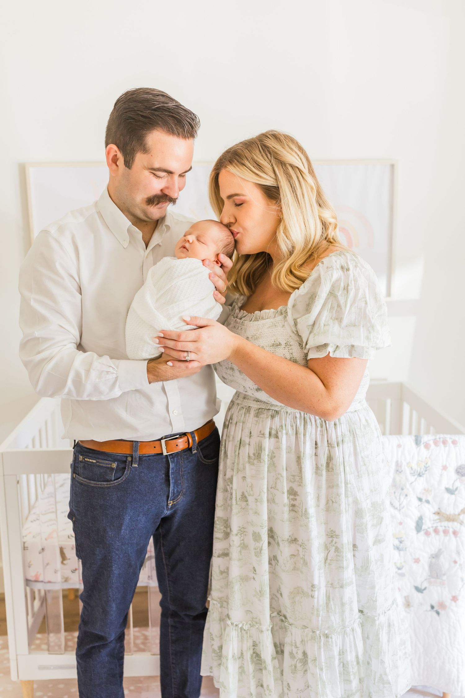 A tender nursery moment with a newborn baby in white being held by parents against a light background.