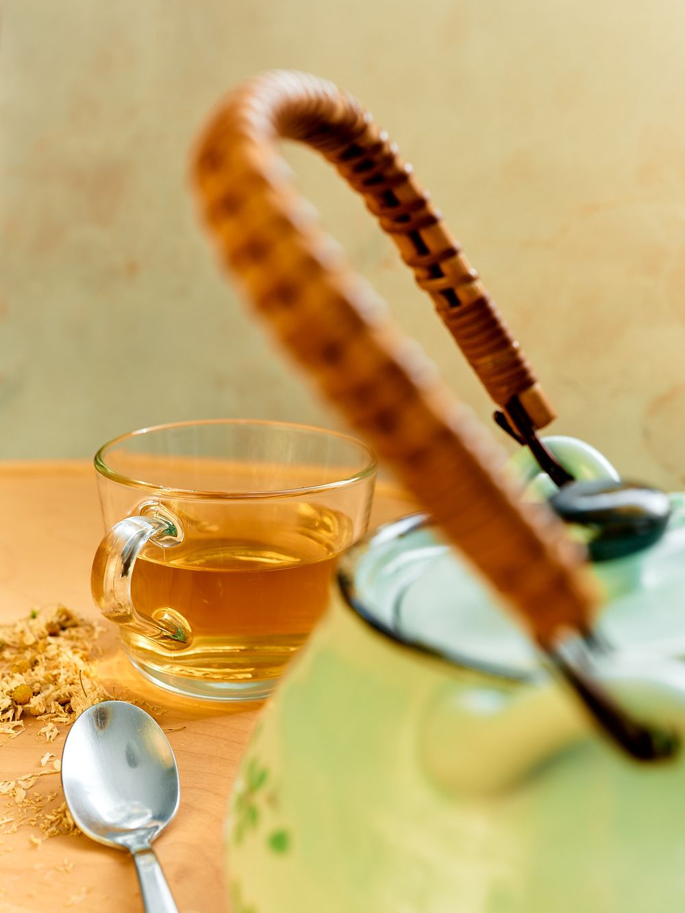 Glass cup of chamomile tea with wooden handled teapot and spoon on pastel background.