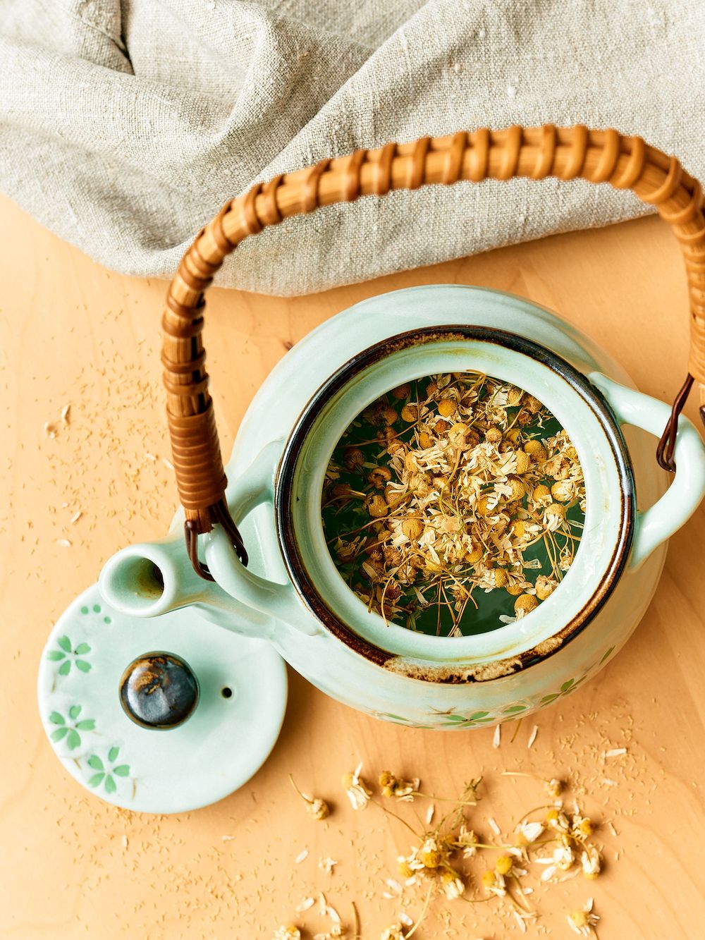 Overhead view of a mint green teapot with chamomile tea leaves and woven bamboo handle.