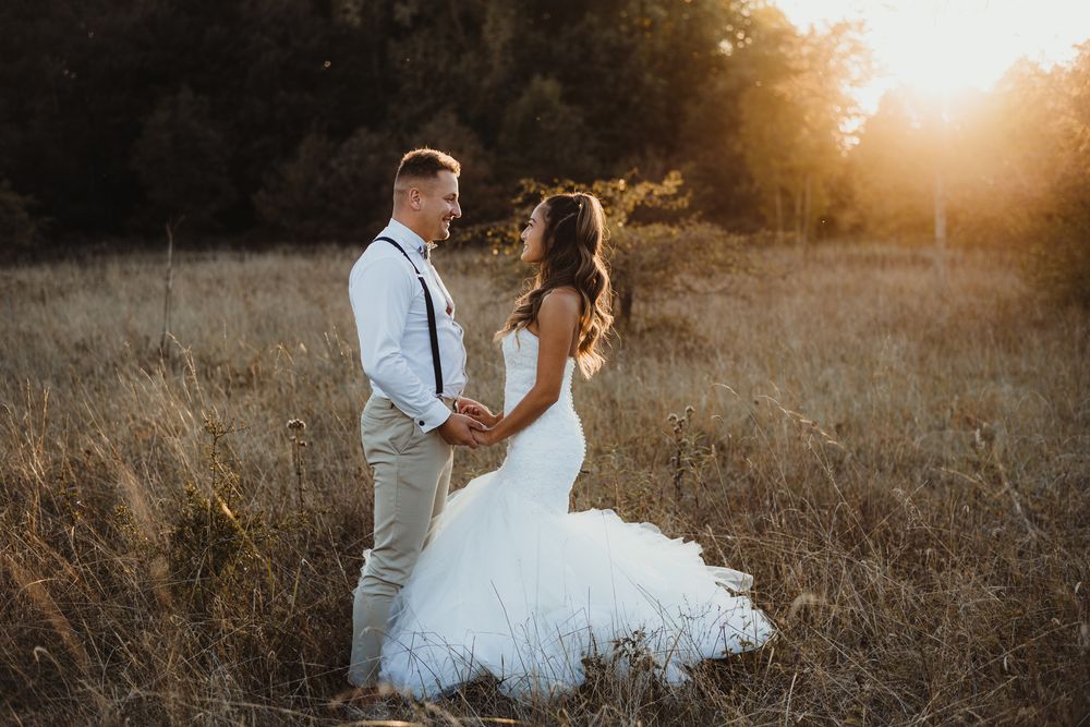 Newlyweds stand together in a sun-lit field wearing classic wedding attire.