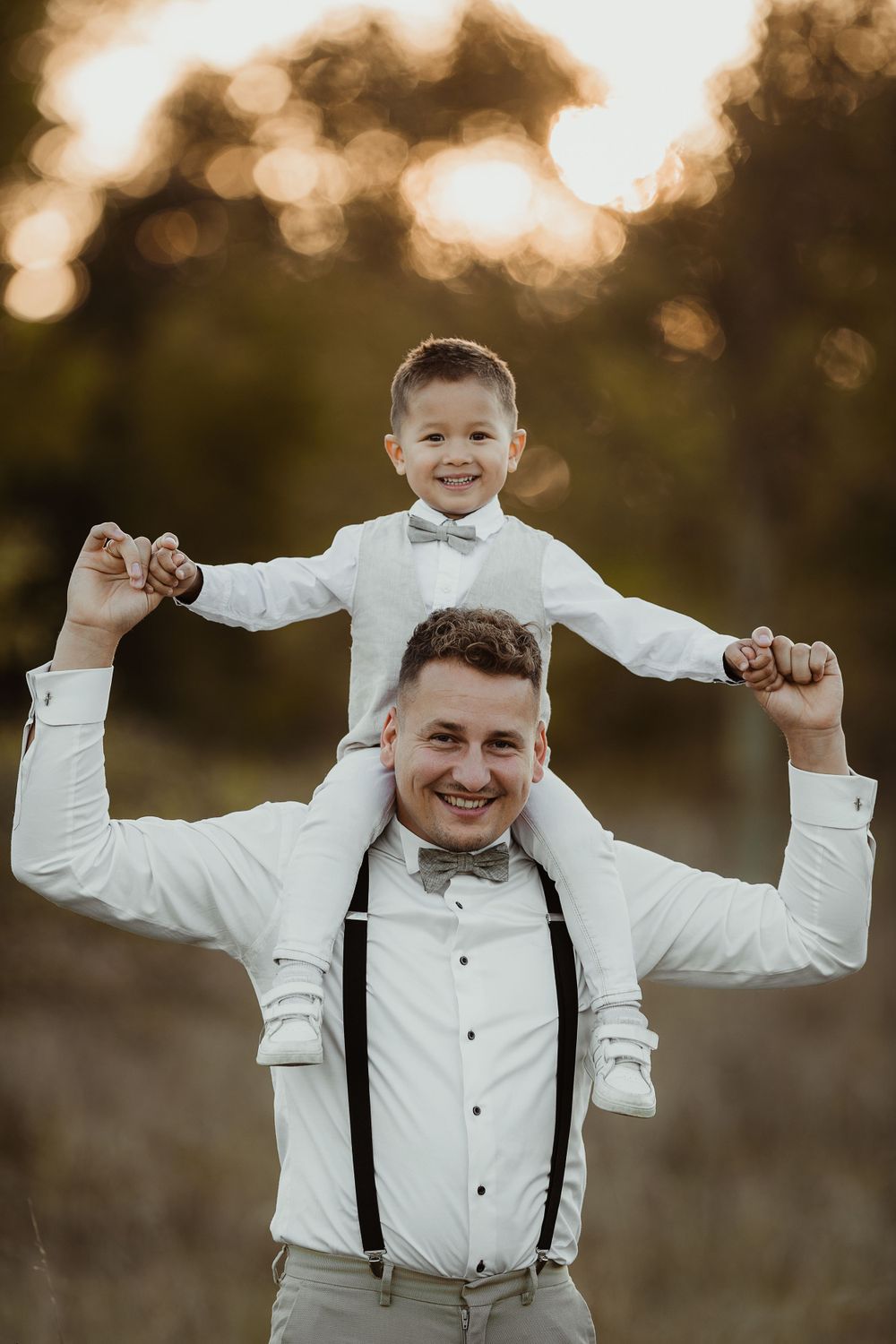 Joyful wedding moment with child riding on shoulders in sunset field.