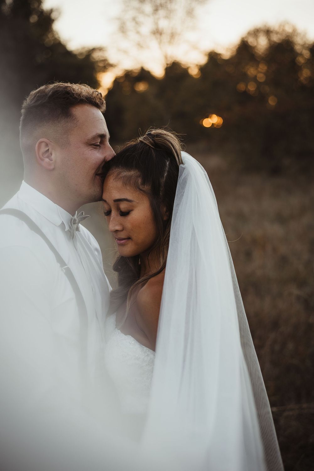 A romantic wedding portrait features a tender moment between newlyweds at sunset with a flowing white veil.