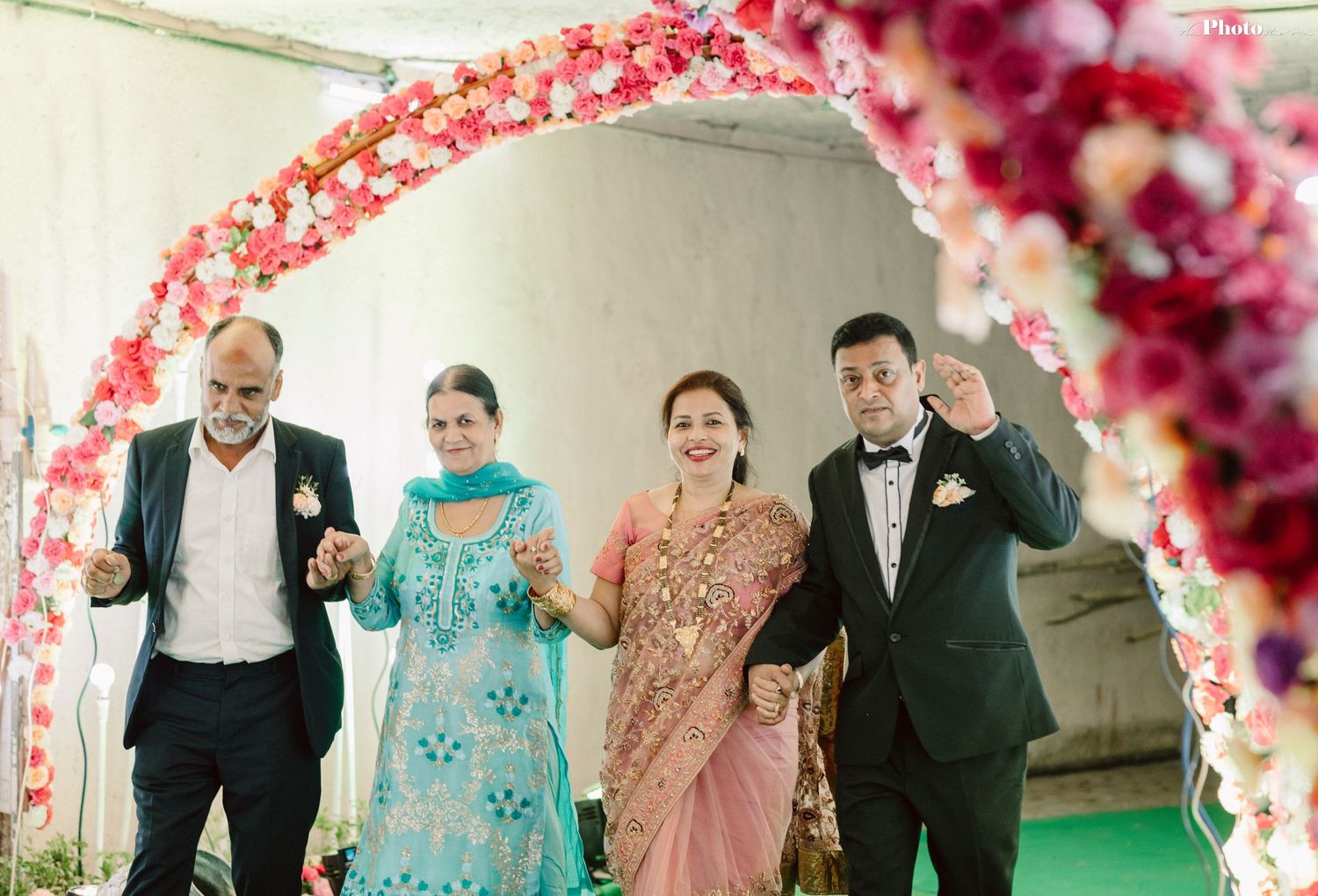 Wedding celebration under floral arch with pink and red blooms and traditional Indian attire.