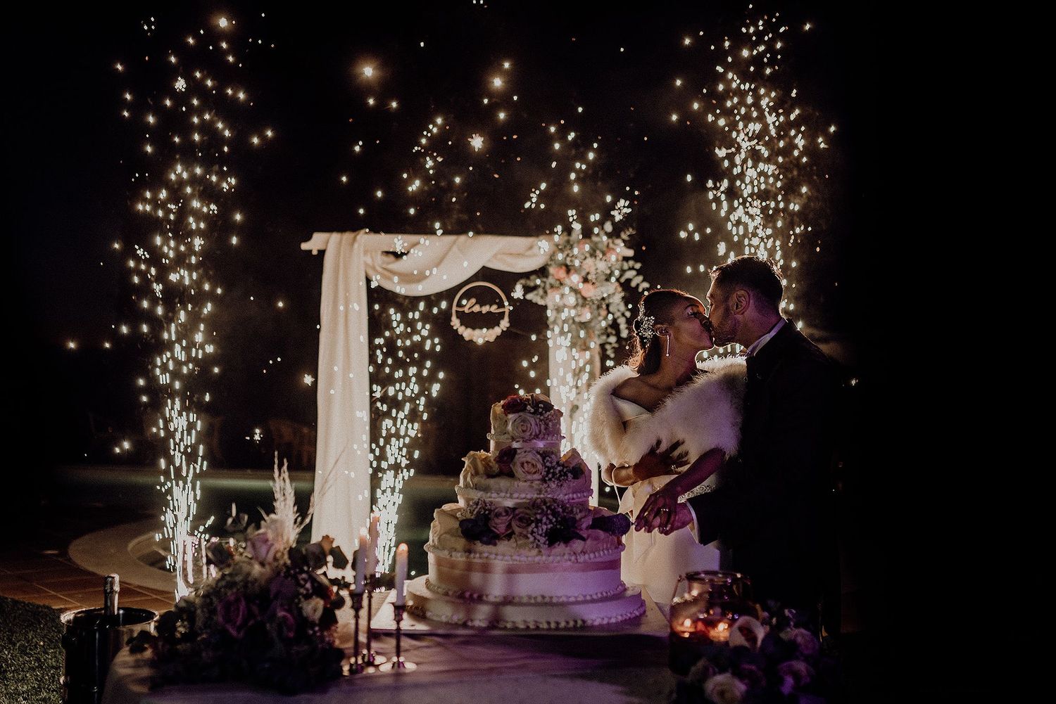 Bride and groom kissing by wedding cake with sparkling fireworks display at romantic night ceremony.