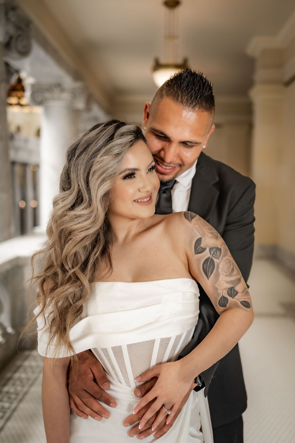 Groom with rose tattoo embraces smiling bride in strapless white wedding dress indoors.