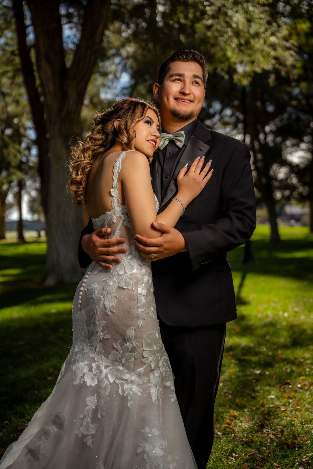 Bride and groom embracing romantically in a sunlit park, wearing wedding gown and black tuxedo.