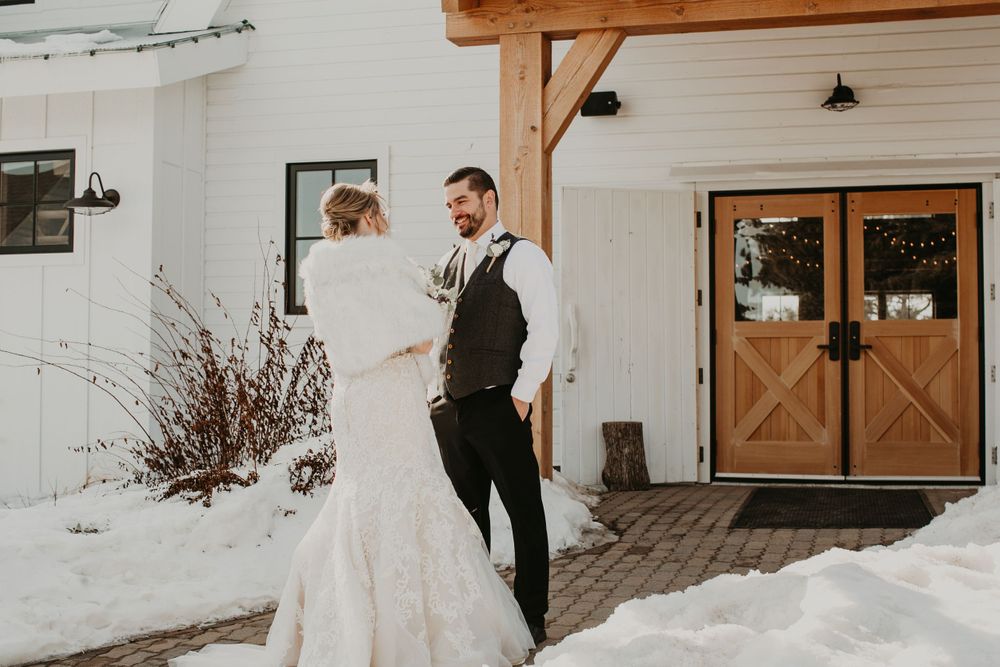 Bride in white lace gown and groom in gray vest share a first look outside a white barn wedding venue in snow.