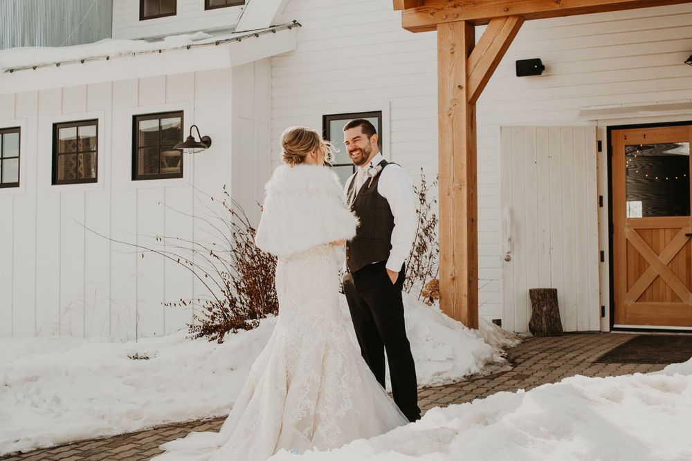 Bride in white lace dress and groom in gray vest share a joyful moment outside a snowy white barn wedding venue.